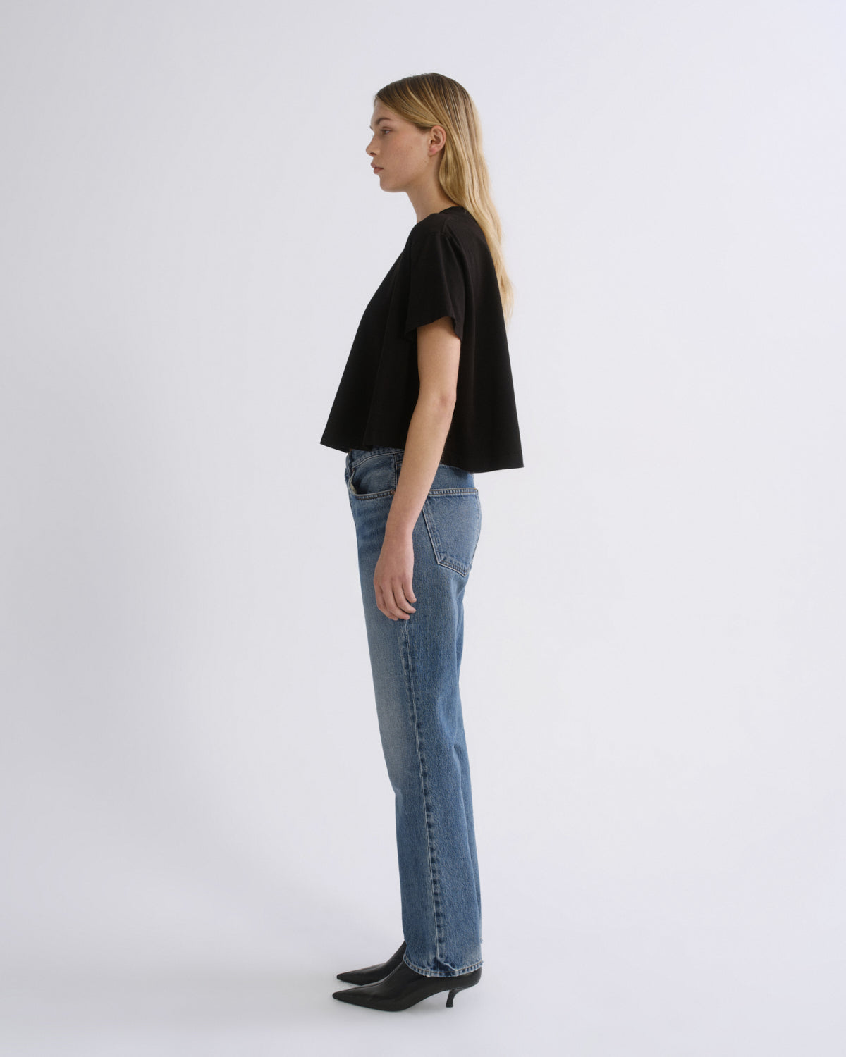 A woman in profile wearing a black cropped short-sleeve top, light-wash blue jeans, and black pointed-toe heels, standing against a plain light gray studio background.