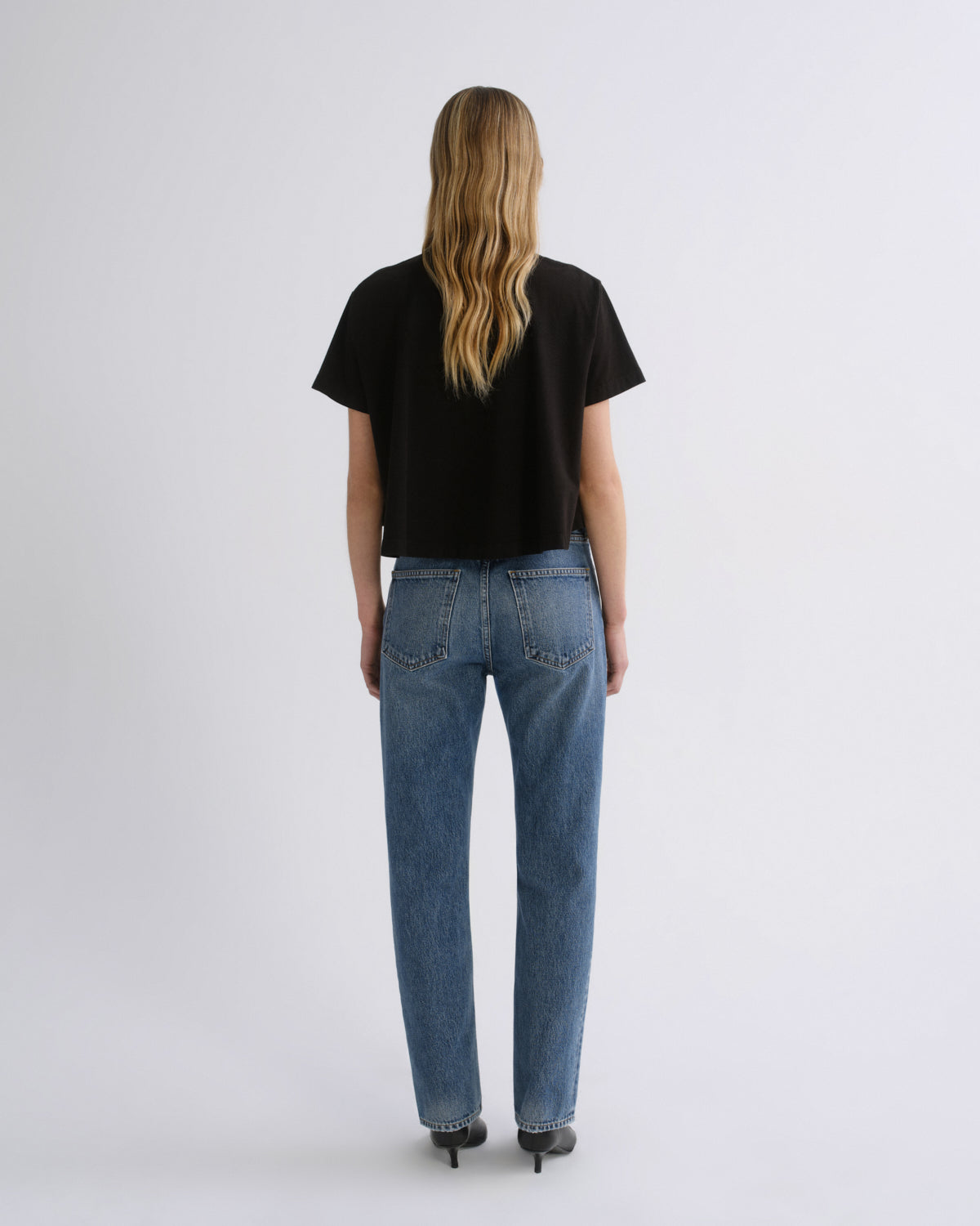 A woman viewed from behind wearing a black short-sleeve t-shirt and blue straight-leg jeans, standing against a plain light gray studio background.