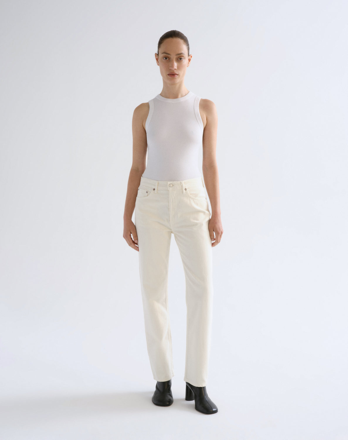 A model stands against a plain light gray studio background wearing a fitted sleeveless white top, off-white straight-leg jeans, and black ankle boots.