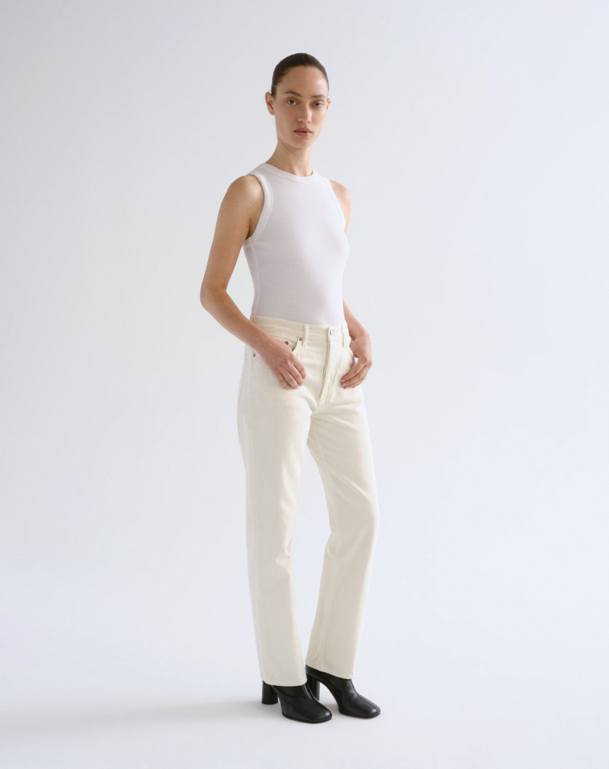 A model stands against a light gray studio background wearing a white sleeveless mock-neck top, off-white straight-leg trousers, and black ankle boots.