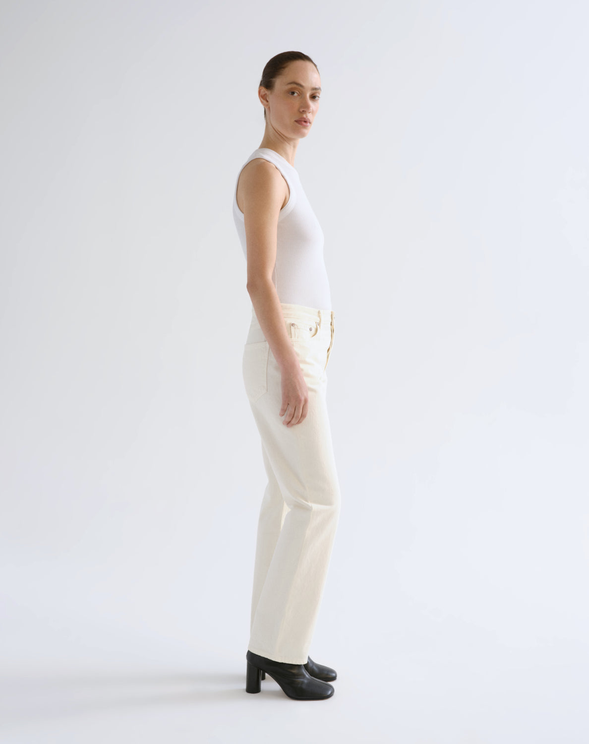 A woman stands in profile wearing a fitted white sleeveless top, off-white straight-leg pants with gold-tone hardware, and black block-heeled shoes against a clean light gray studio background.
