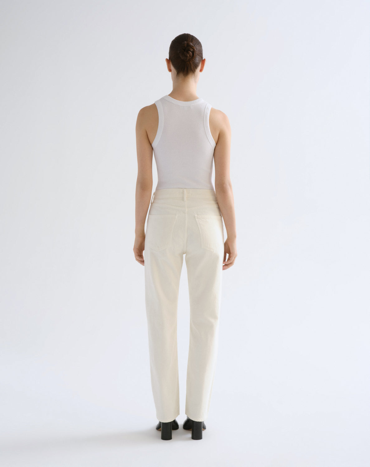 A model shown from the back wearing a white sleeveless top and off-white straight-leg trousers, standing against a plain light gray studio background.