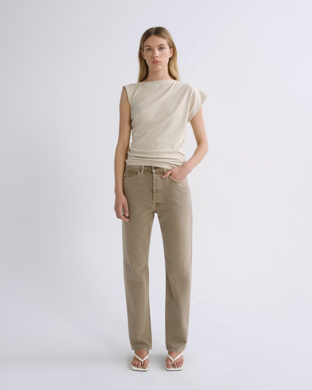 A woman stands against a light gray studio background wearing a sleeveless cream top and straight-leg beige jeans, with white sandals.