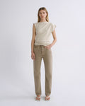 A woman stands against a light gray studio background wearing a sleeveless cream top and straight-leg beige jeans, with white sandals.
