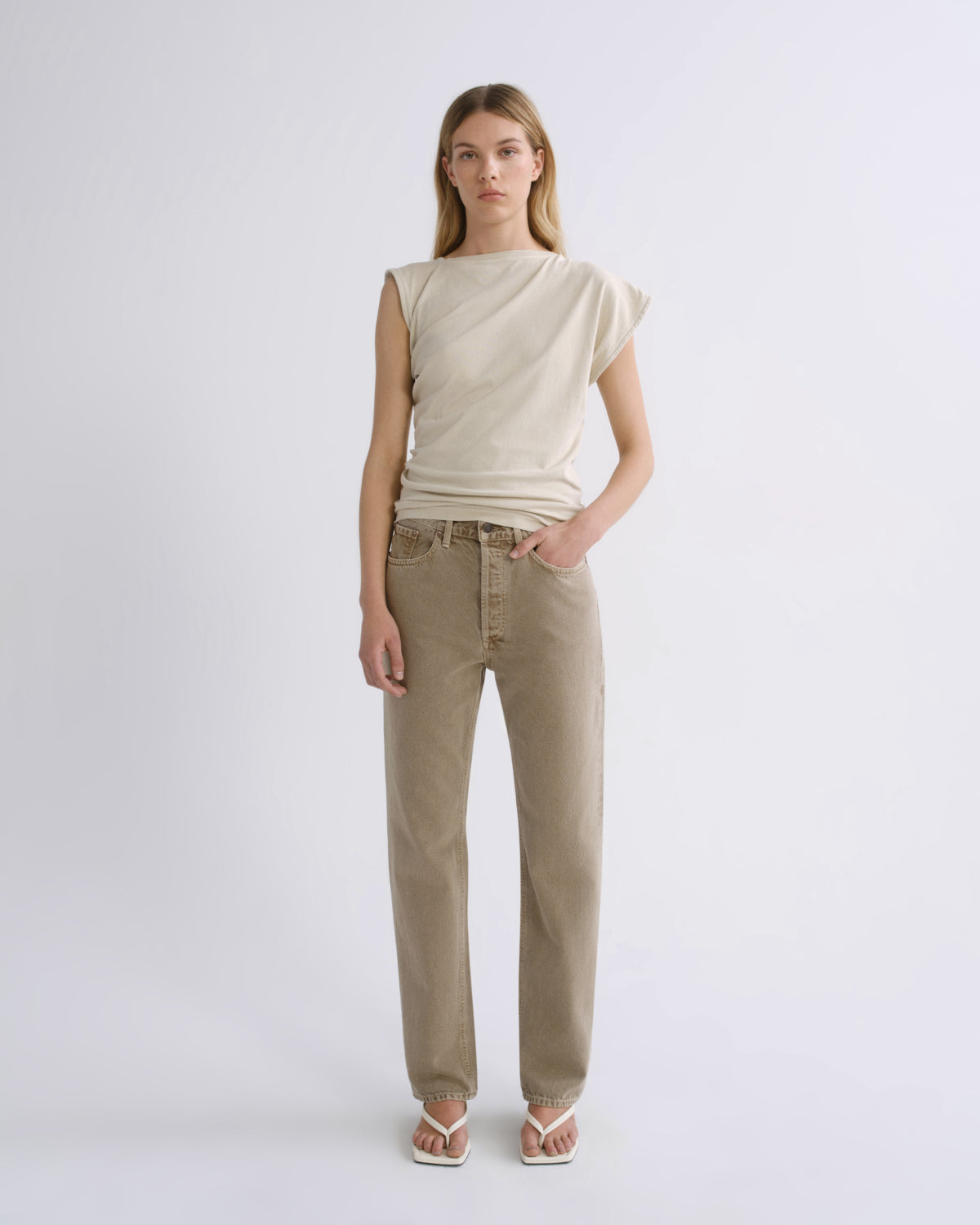 A woman stands against a light gray studio background wearing a sleeveless cream top and straight-leg beige jeans, with white sandals.