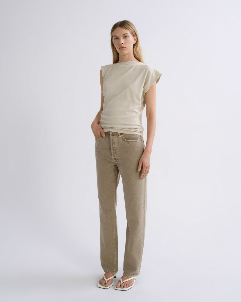 A woman modeling a light beige short-sleeve top and beige straight-leg jeans against a plain light gray studio background.