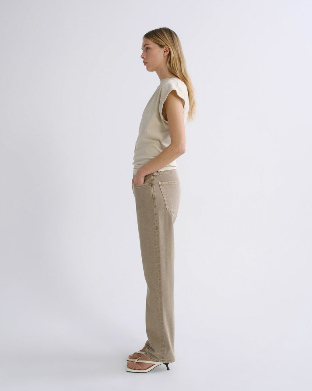 A woman stands in profile wearing a sleeveless cream top and beige wide-leg pants against a plain light gray studio background.