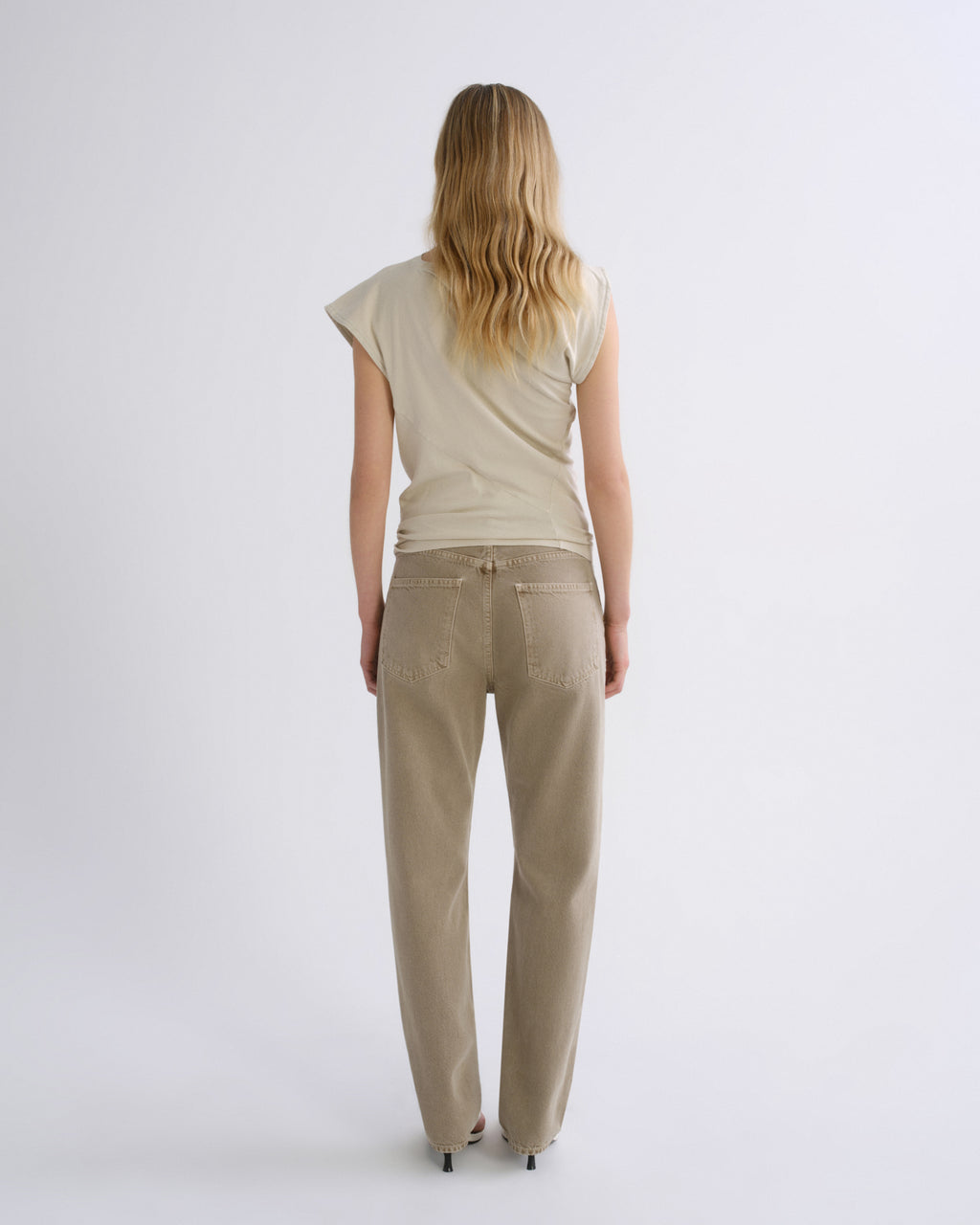 A woman viewed from behind wearing a light beige short-sleeve top and matching beige straight-leg pants against a plain light gray studio background.
