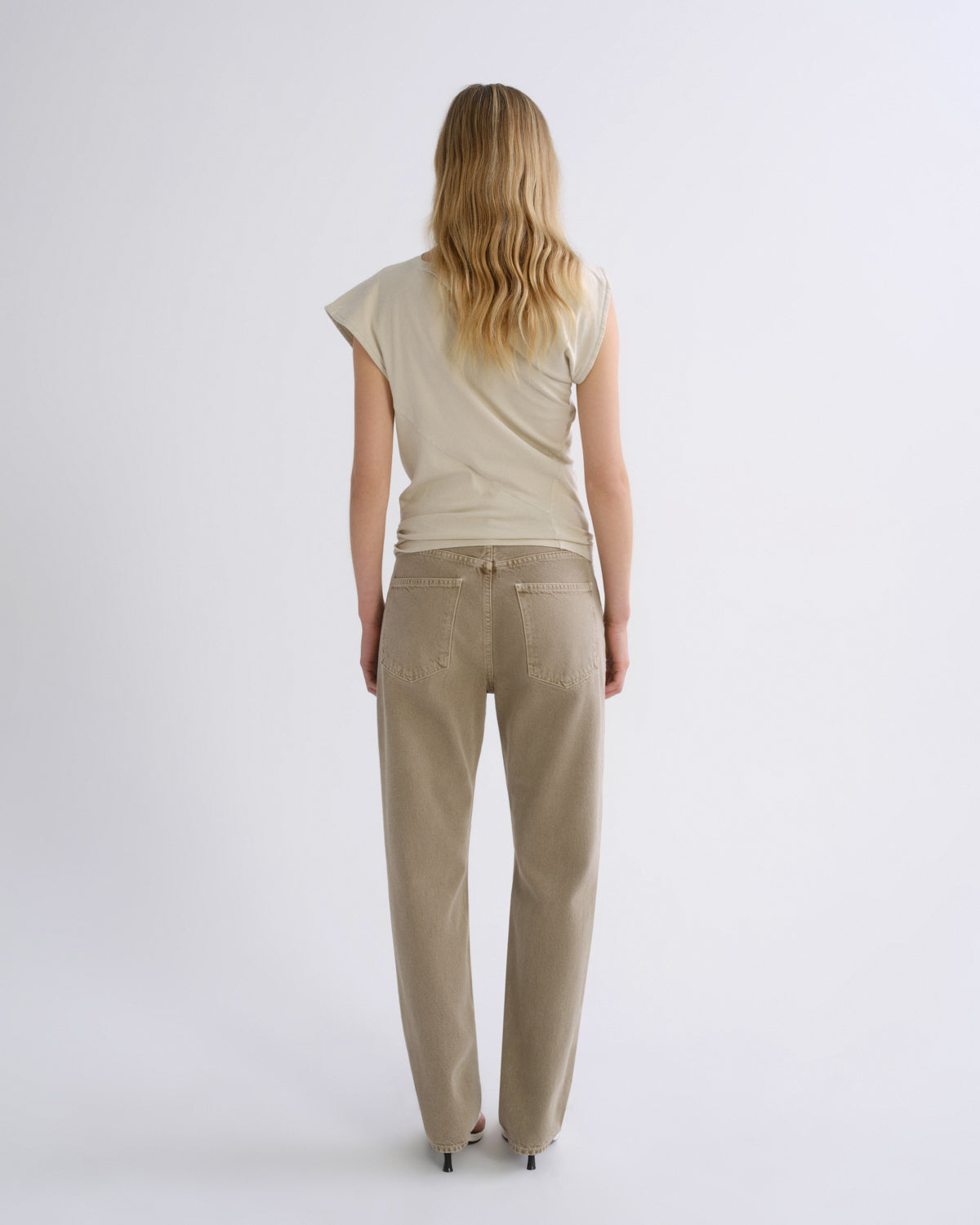 A woman viewed from behind wearing a light beige short-sleeve top and matching beige straight-leg pants against a plain light gray studio background.