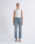 A model stands against a light gray studio background wearing a white ribbed sleeveless top and light-wash blue straight-leg jeans with beige strappy heels.