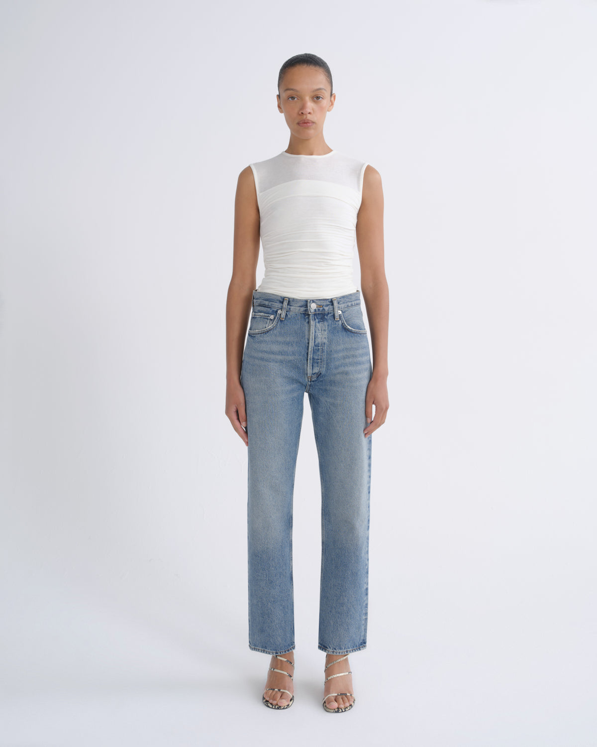 A model stands against a light gray studio background wearing a white ribbed sleeveless top and light-wash blue straight-leg jeans with beige strappy heels.
