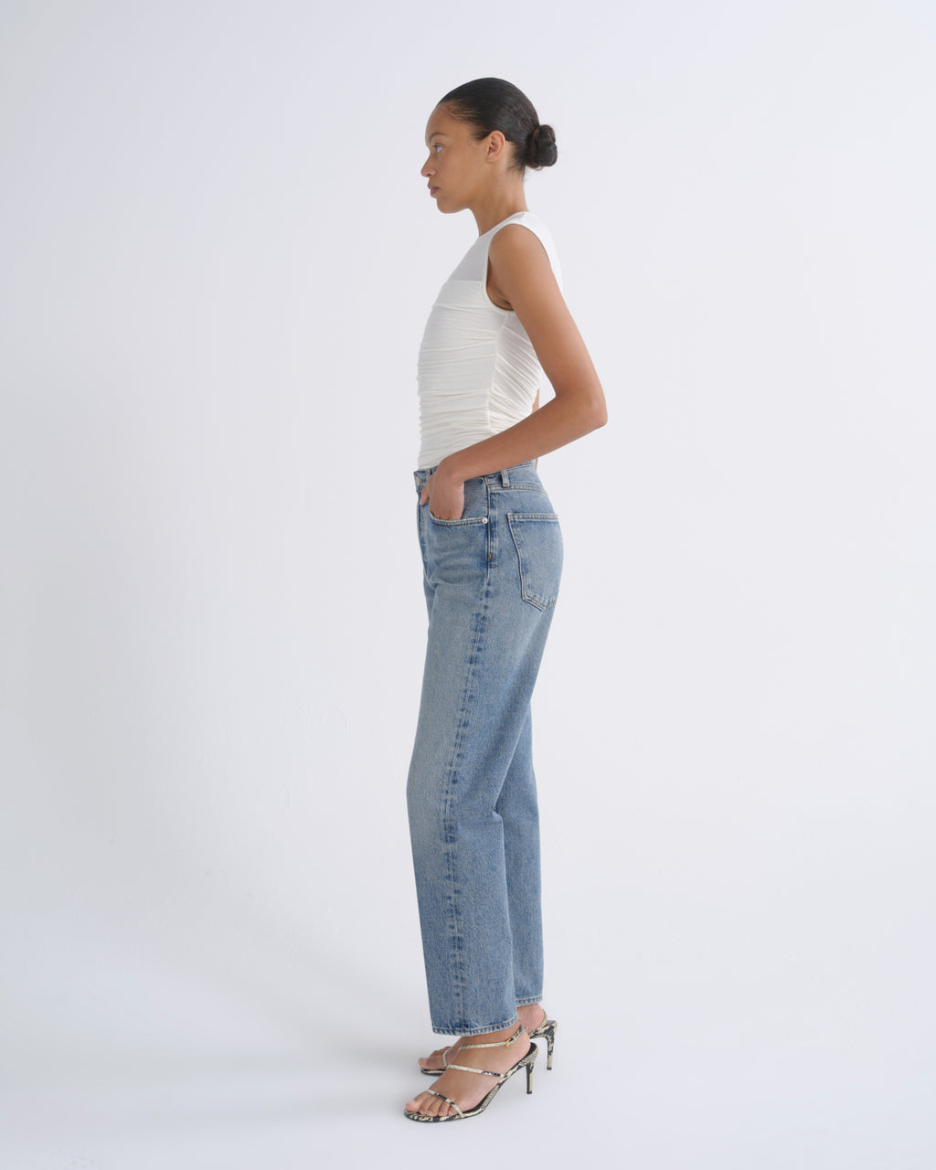 A woman in profile wearing light-wash blue jeans, a white ribbed sleeveless top, and metallic heeled sandals, standing against a clean white studio background.