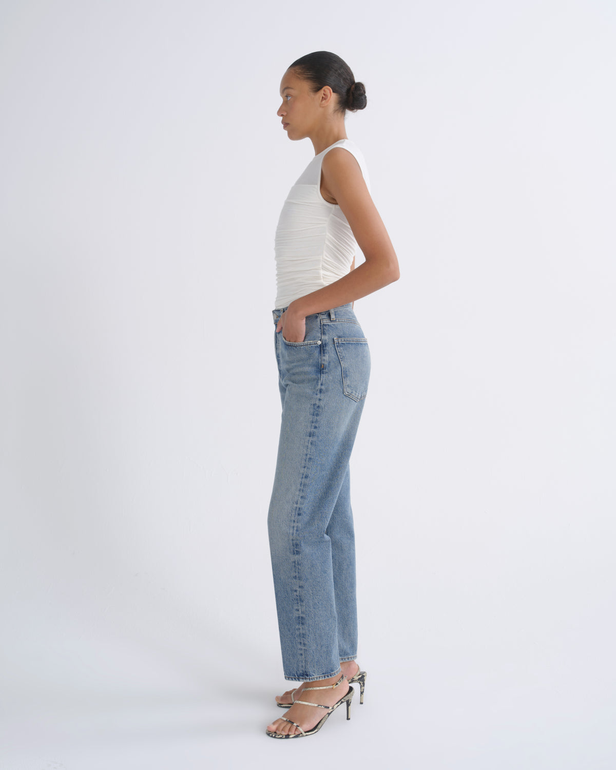 A woman in profile wearing light-wash blue jeans, a white ribbed sleeveless top, and metallic heeled sandals, standing against a clean white studio background.