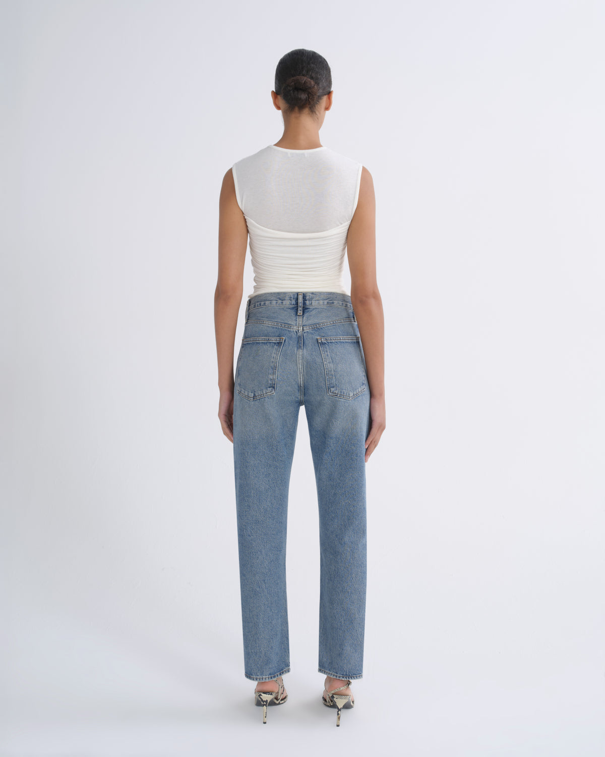 A model viewed from the back wearing a white ribbed sleeveless top and light-wash blue straight-leg jeans, paired with metallic high-heeled sandals, against a plain light gray studio background.