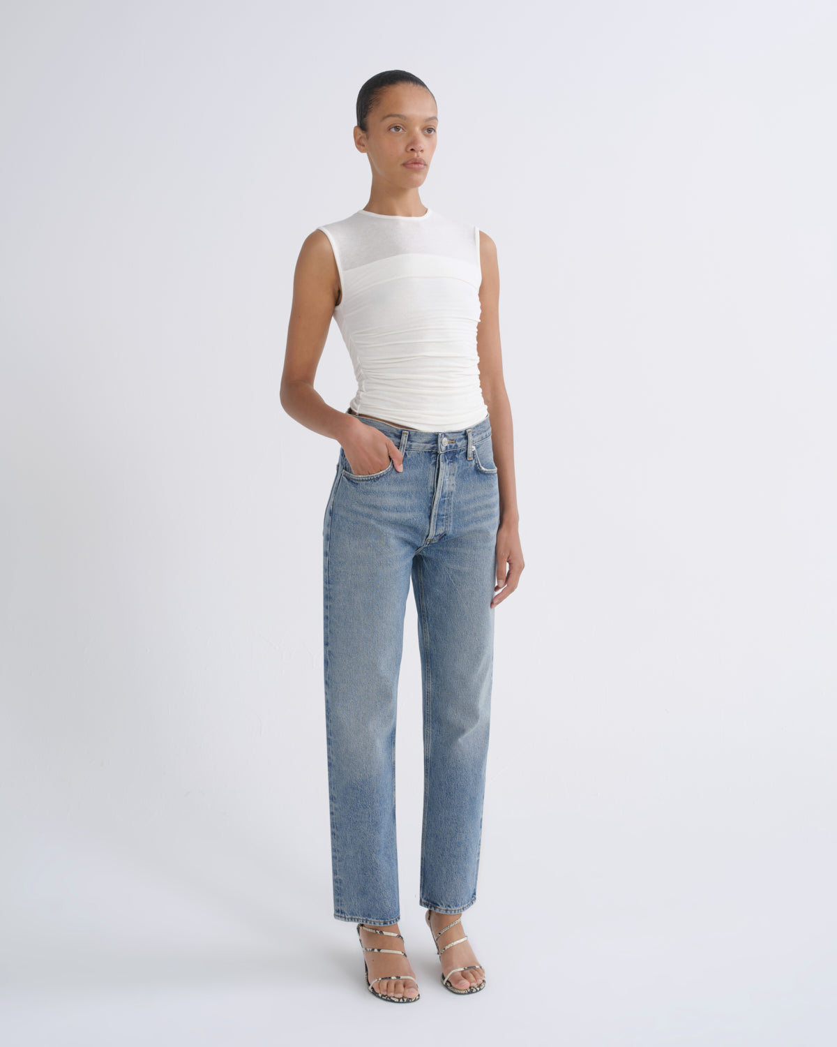 A model stands against a light gray studio background wearing a sleeveless white ribbed top and light-wash blue straight-leg jeans with beige strappy heels.