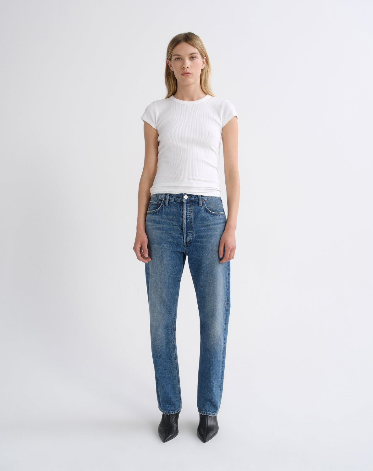 A woman stands against a light gray studio background wearing a fitted white short-sleeve top, blue straight-leg jeans, and black pointed ankle boots.