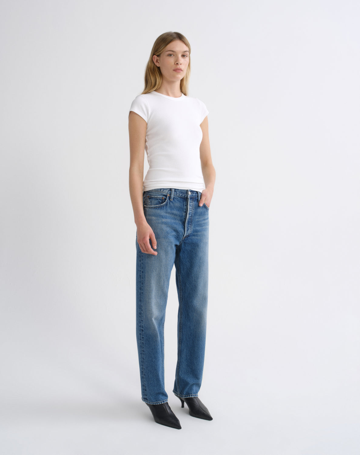 A woman modeling a white short-sleeve crewneck t-shirt and straight-leg blue jeans with black pointed-toe heels, standing against a light gray studio background.