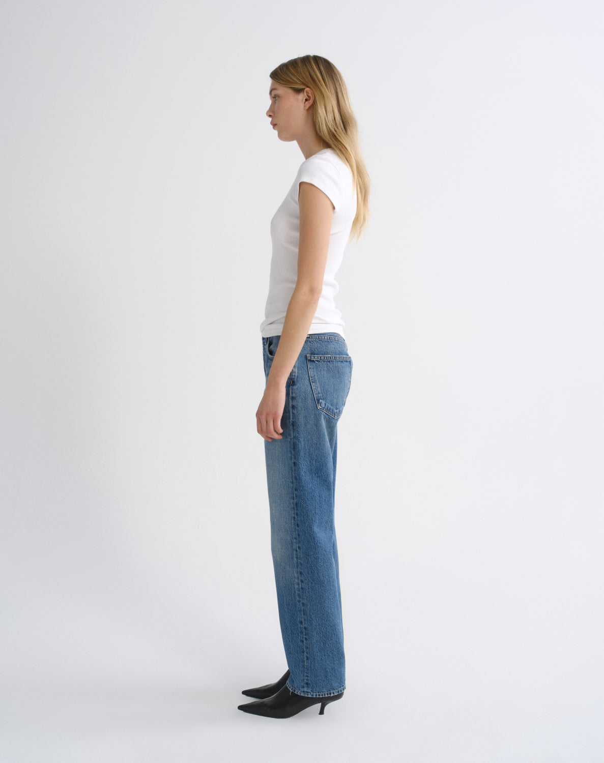 A woman stands in profile wearing a white short-sleeve top, blue straight-leg jeans, and black pointed-toe heels against a clean white studio background.