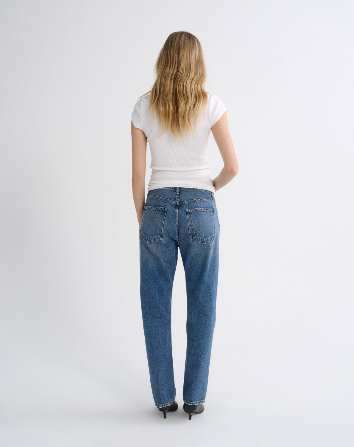 A woman viewed from behind wearing blue straight-leg jeans, a white short-sleeve t-shirt, and black heels against a light gray studio background.