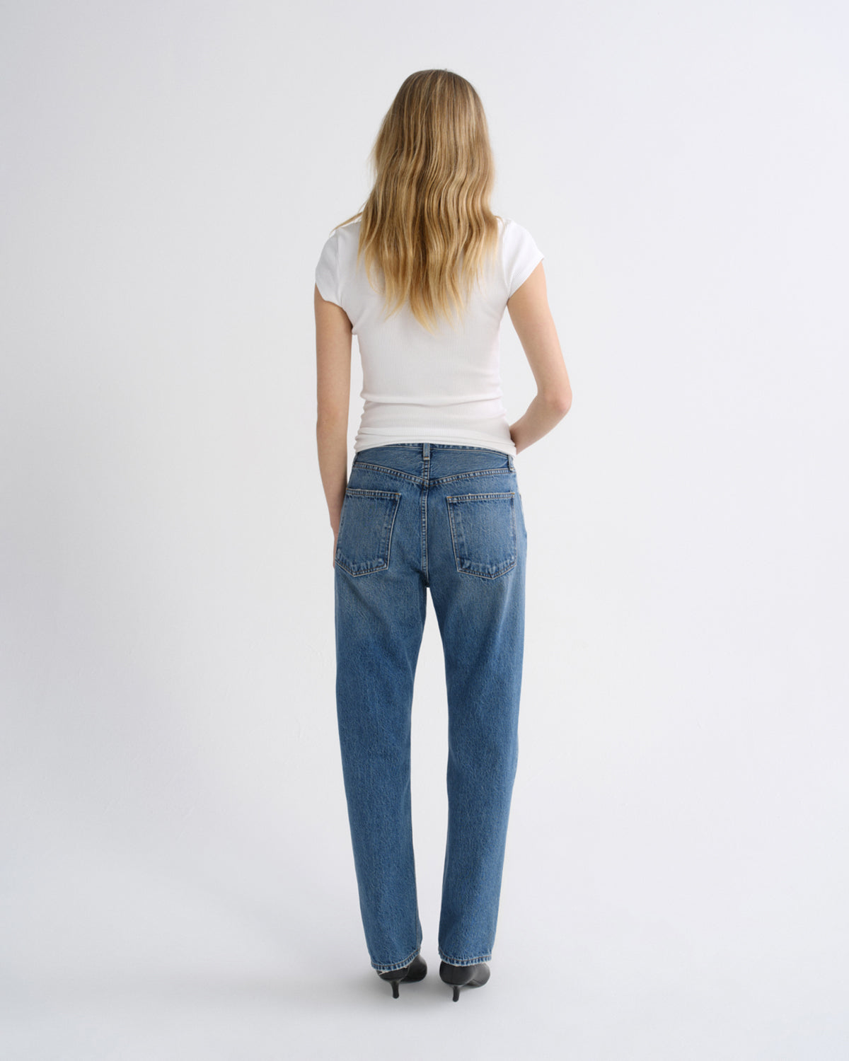 A woman stands against a light gray studio background wearing a fitted white short-sleeve top, blue straight-leg jeans, and black pointed ankle boots.