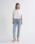 A model stands against a light gray studio background wearing a white short-sleeve cropped T-shirt, light-wash blue straight-leg jeans, and beige strappy high-heeled sandals.
