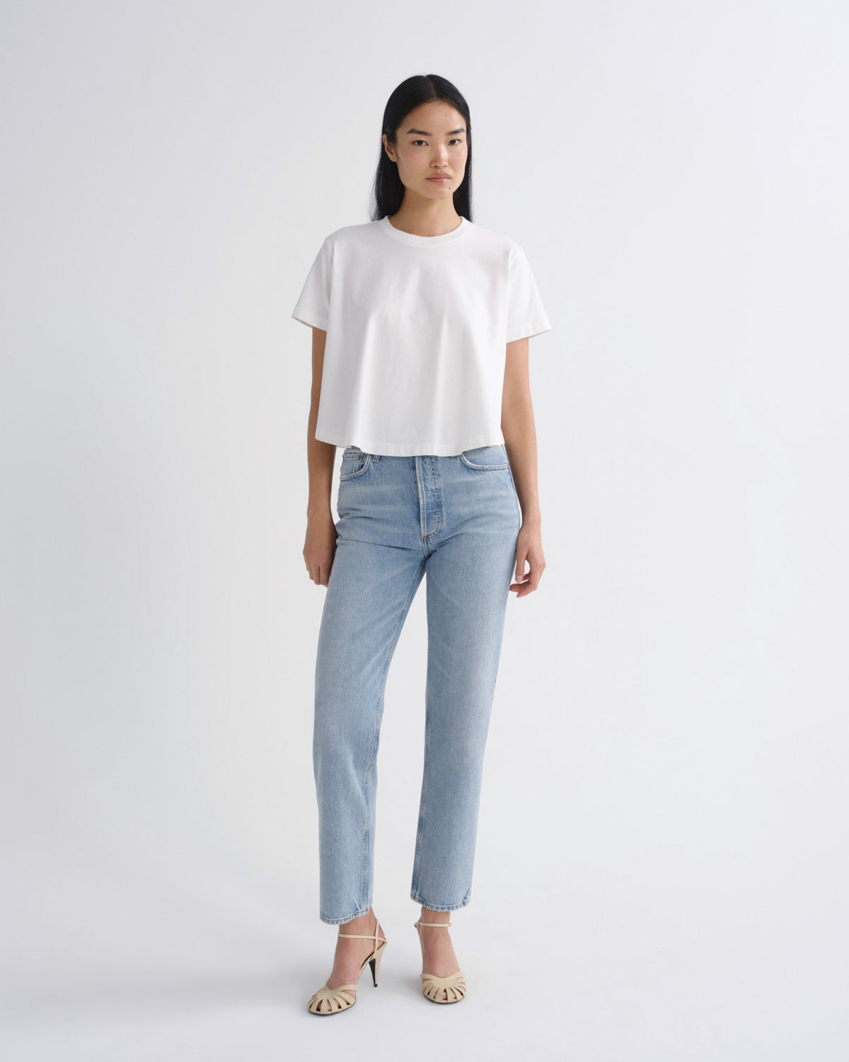 A model stands against a light gray studio background wearing a white short-sleeve cropped T-shirt, light-wash blue straight-leg jeans, and beige strappy high-heeled sandals.