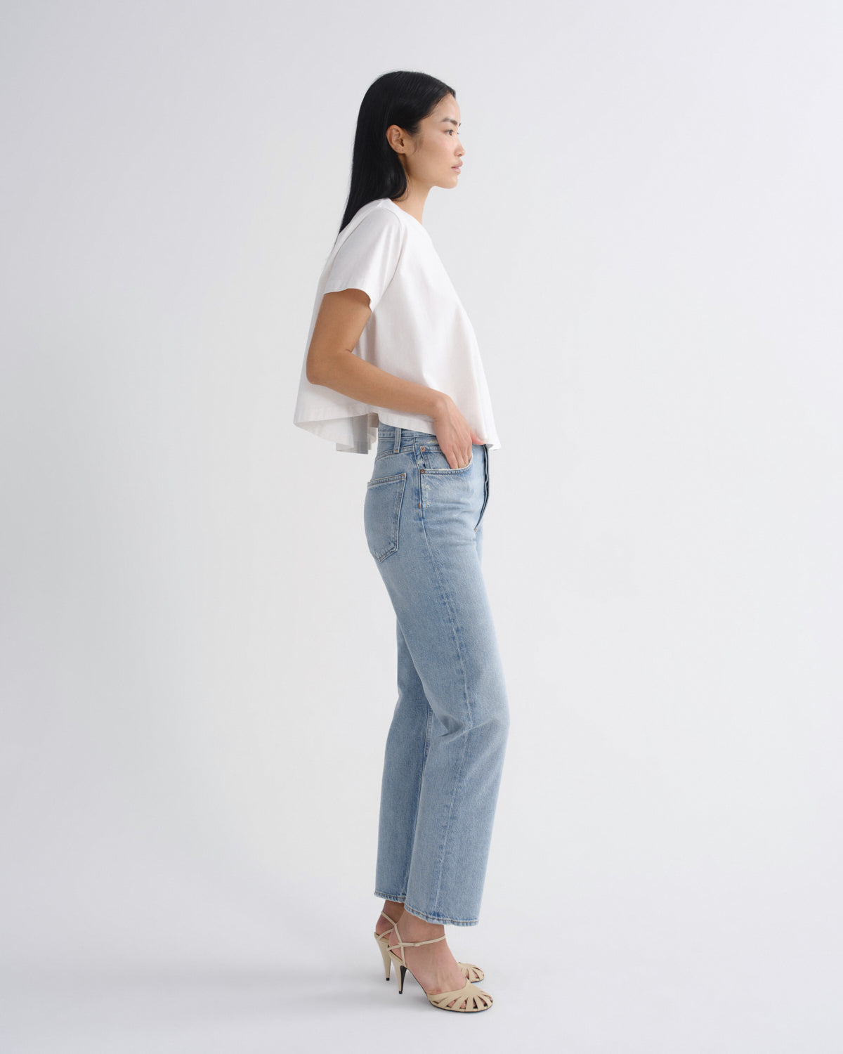 A woman in profile wearing a white short-sleeve top, light-wash blue jeans, and beige high-heeled sandals, standing against a plain light gray studio background.