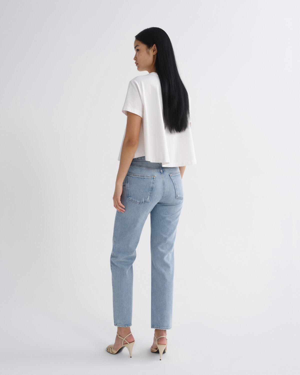 A woman is shown from the back wearing a white short-sleeve top, light-wash blue jeans, and beige high-heeled sandals against a plain white studio background.