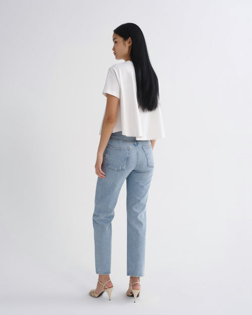 A woman is shown from the back wearing a white short-sleeve top, light-wash blue jeans, and beige high-heeled sandals against a plain white studio background.