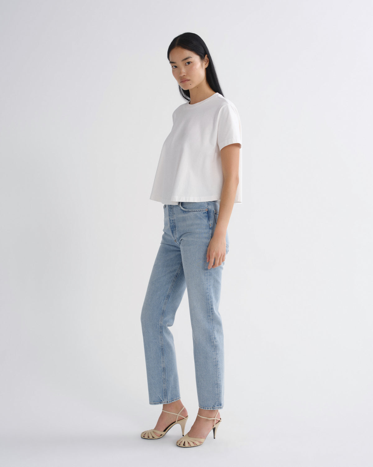 A woman modeling a white short-sleeve crop top and light-wash blue jeans with beige strappy heels, standing against a plain light gray studio background.