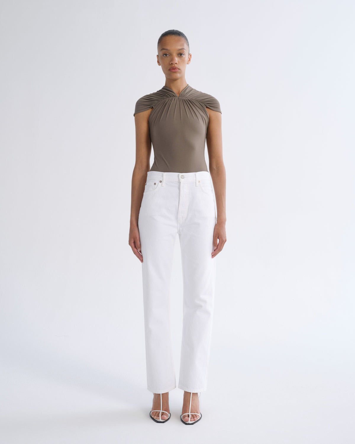 A model stands against a light gray studio background wearing a taupe-brown draped short-sleeve top and white straight-leg jeans with clear-strap heeled sandals.