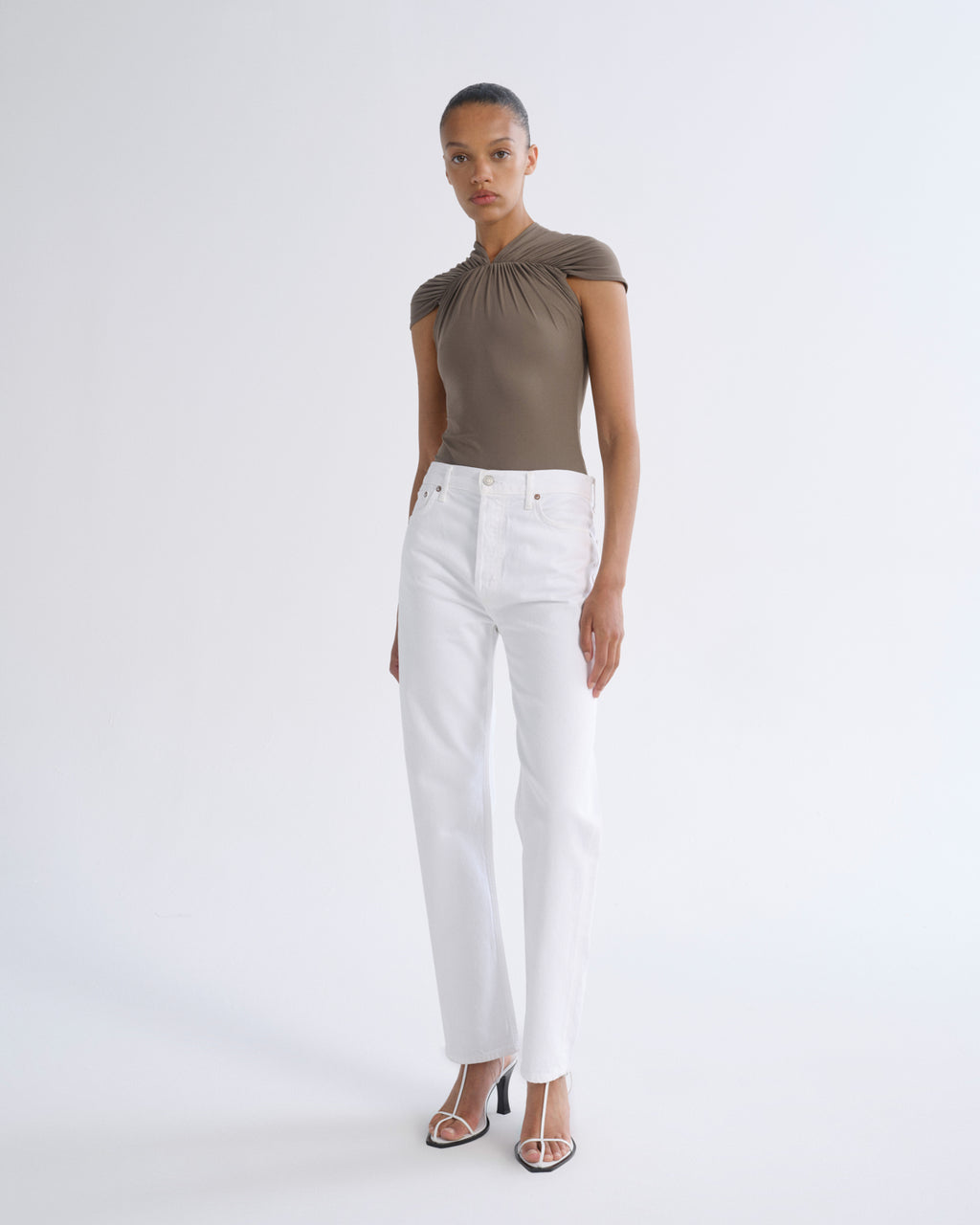A model stands against a light gray studio background wearing a taupe-brown one-shoulder top, white straight-leg jeans, and clear high-heeled sandals.