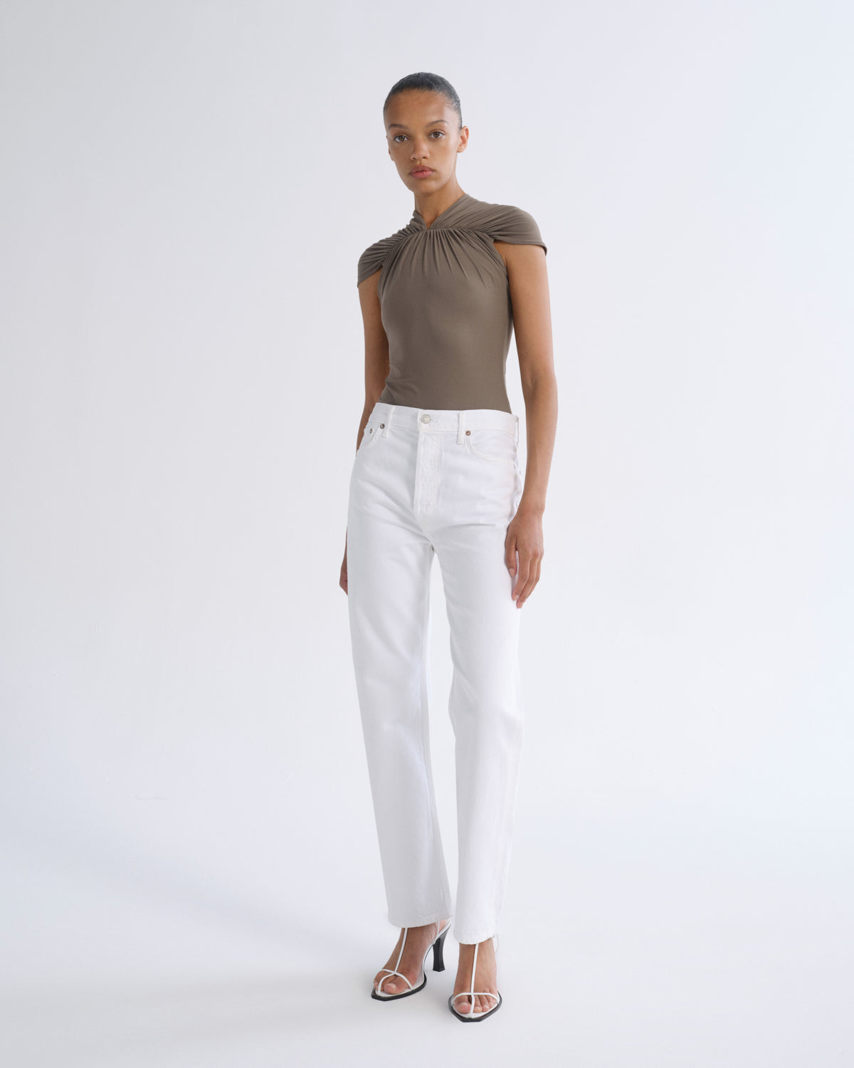 A model stands against a light gray studio background wearing a taupe-brown one-shoulder top, white straight-leg jeans, and clear high-heeled sandals.