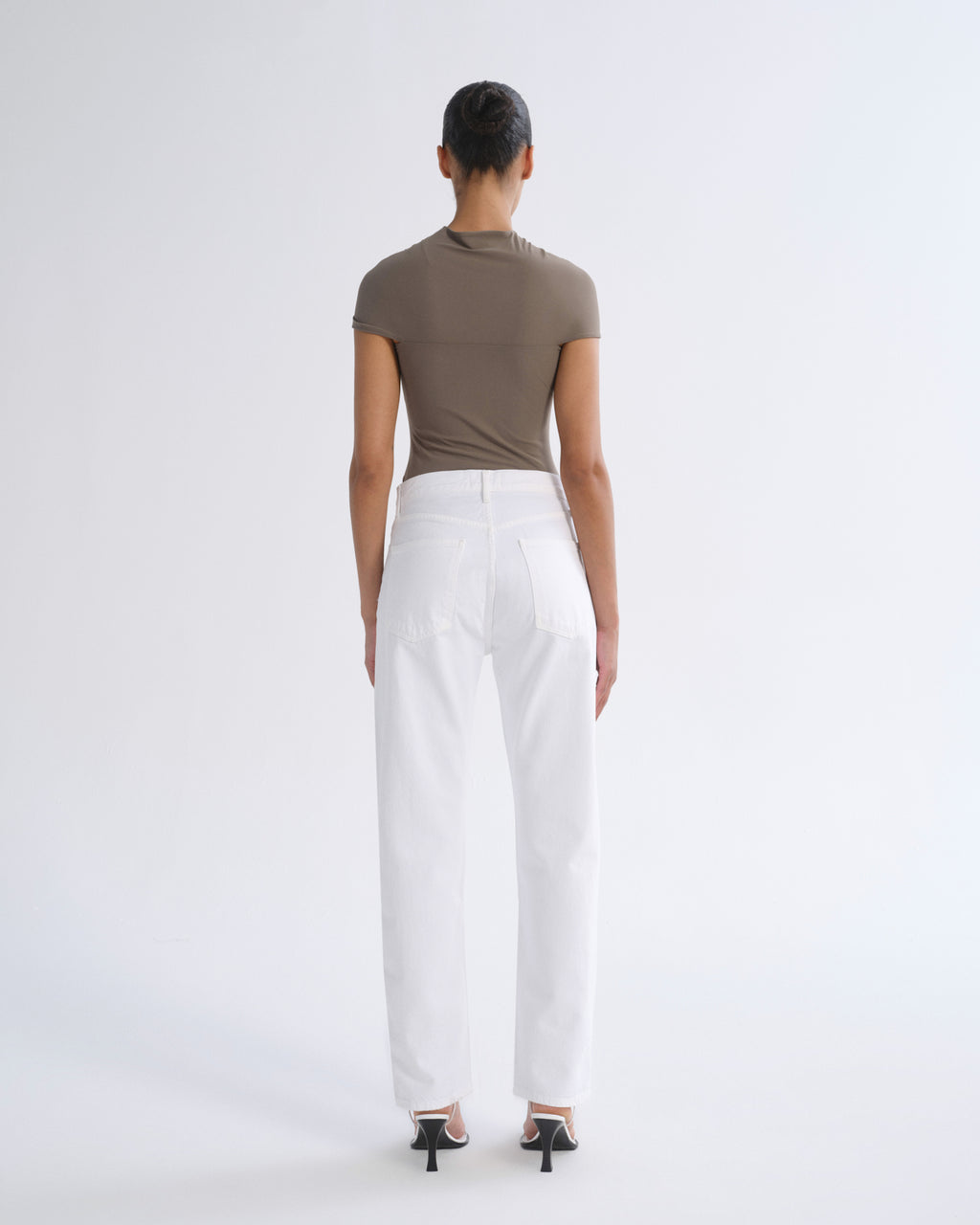 A model shown from the back wearing a fitted taupe short-sleeve top and white wide-leg trousers, paired with black high-heeled sandals, against a plain light gray studio background.