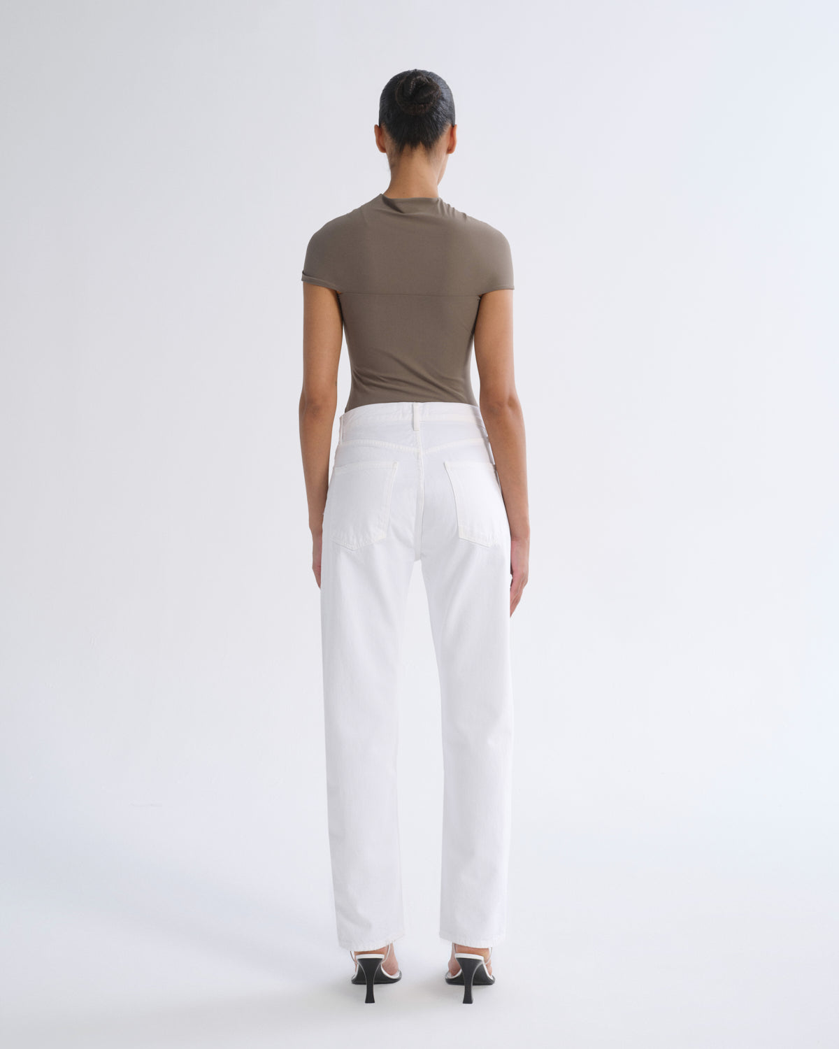 A model shown from the back wearing a fitted taupe short-sleeve top and white wide-leg trousers, paired with black high-heeled sandals, against a plain light gray studio background.