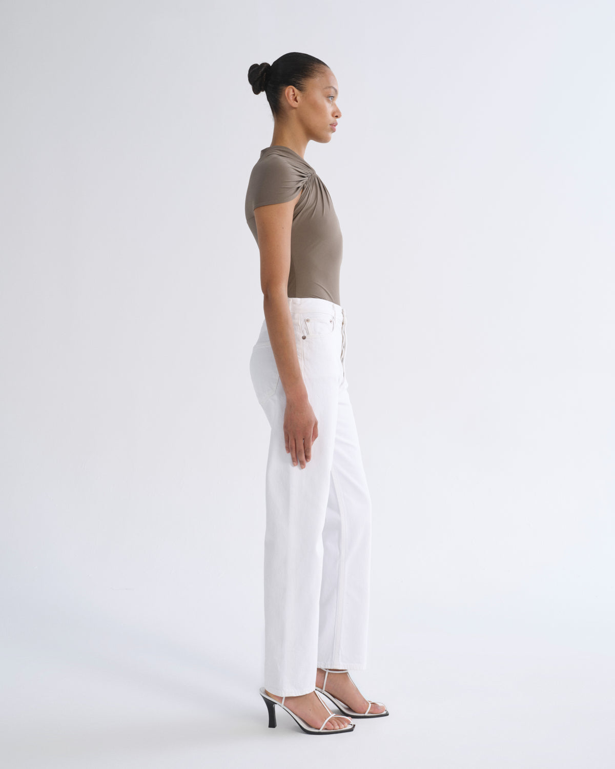 A woman in profile wearing a taupe short-sleeve ruched top, white wide-leg cropped pants, and silver heeled sandals, standing against a light gray studio background.