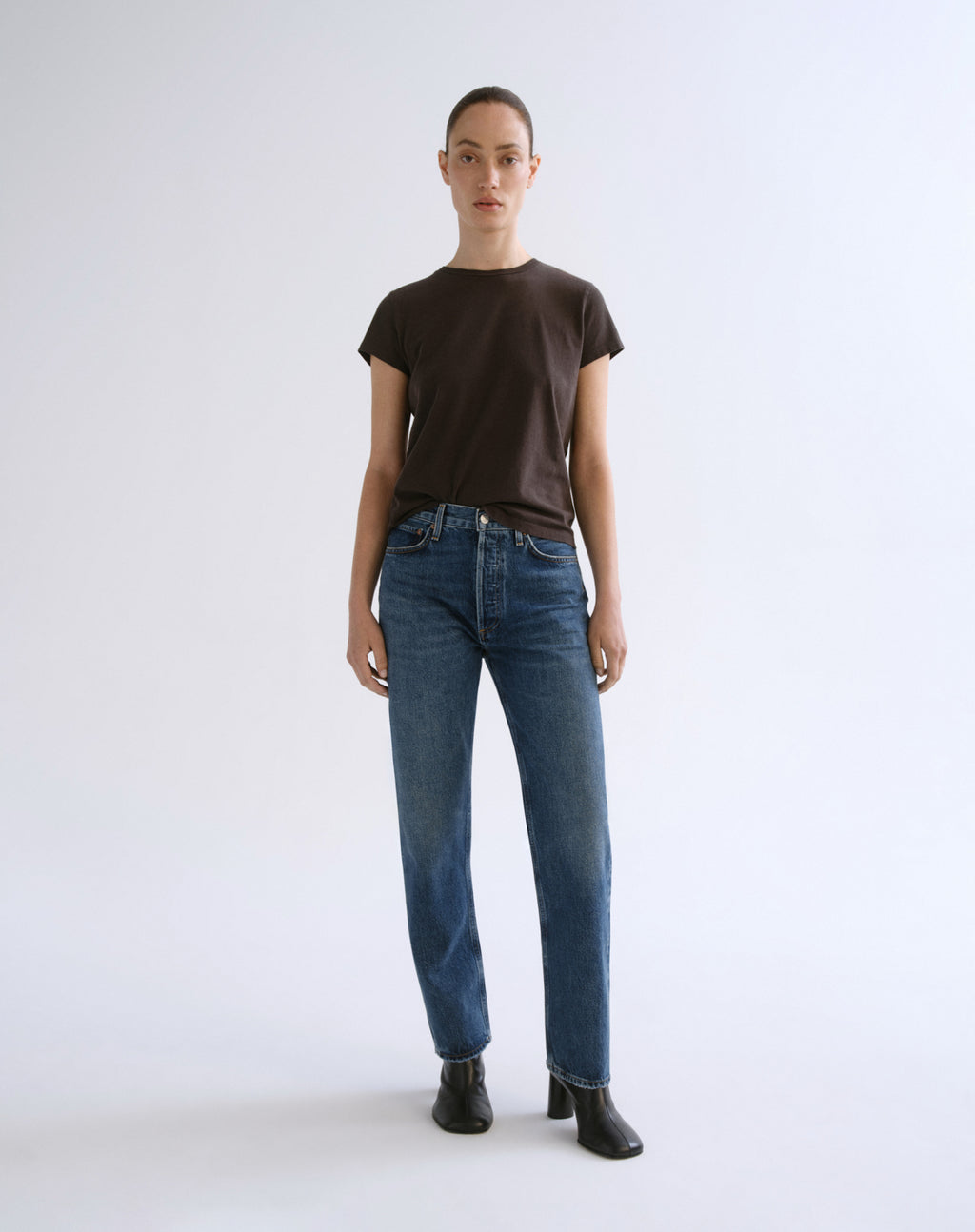 A model stands against a light gray studio background wearing a dark brown short-sleeve crewneck t-shirt, blue straight-leg jeans, and black ankle boots.