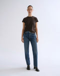 A model stands against a light gray studio background wearing a dark brown short-sleeve crewneck t-shirt, blue straight-leg jeans, and black ankle boots.