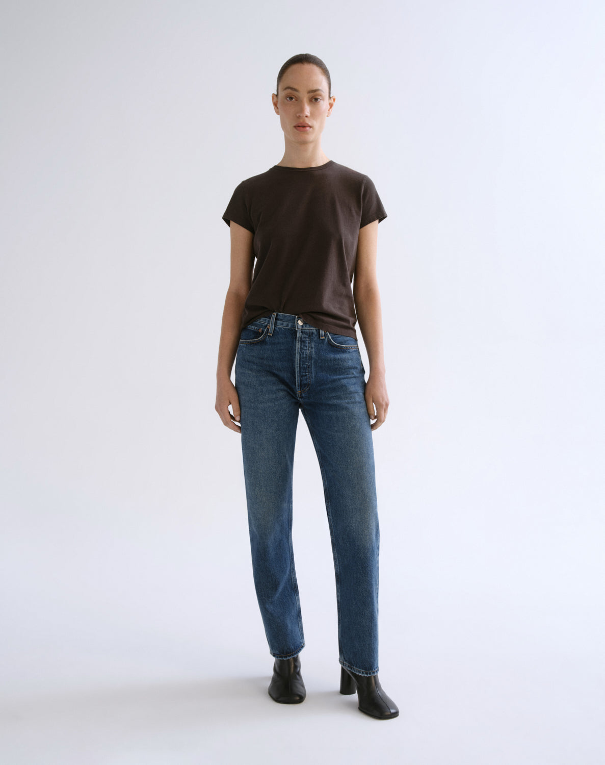 A model stands against a light gray studio background wearing a dark brown short-sleeve crewneck t-shirt, blue straight-leg jeans, and black ankle boots.