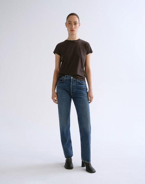 A model stands against a light gray studio background wearing a dark brown short-sleeve crewneck t-shirt, blue straight-leg jeans, and black ankle boots.