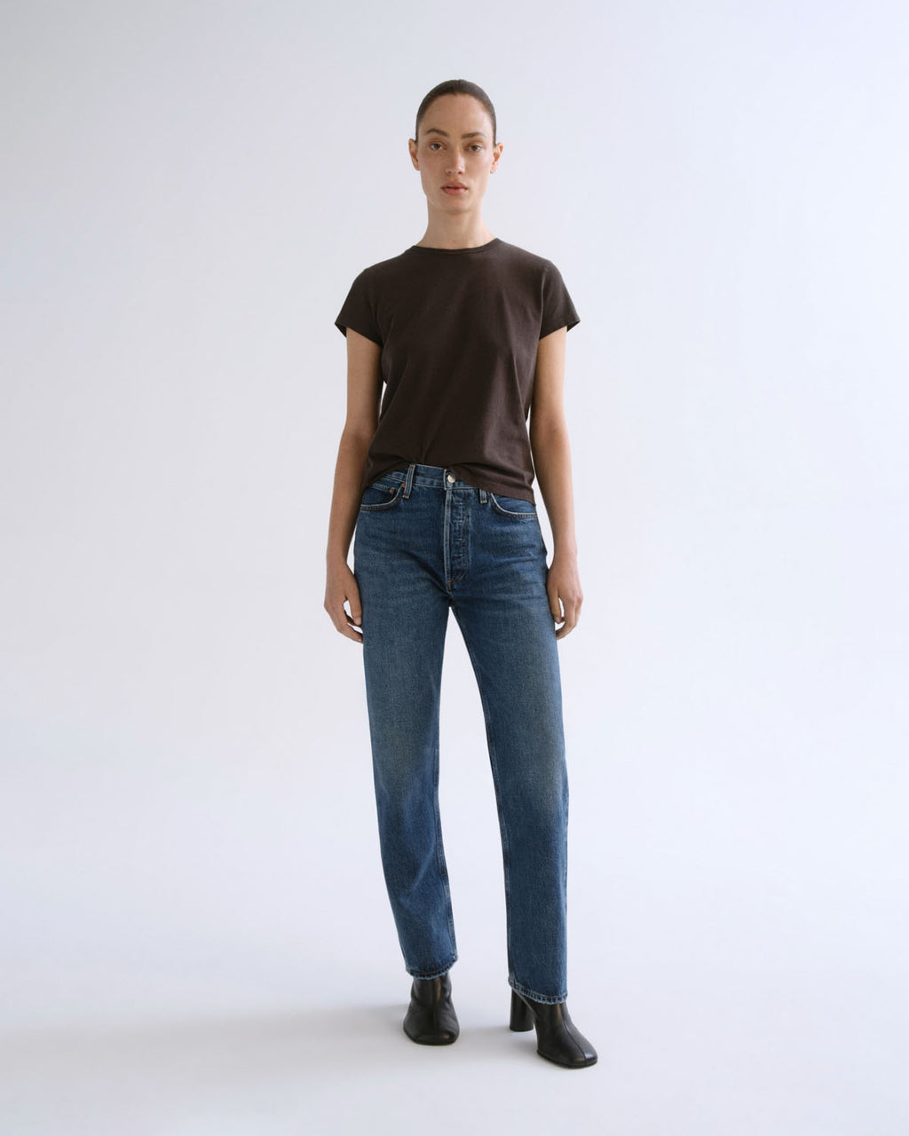 A model stands against a light gray studio background wearing a dark brown short-sleeve crewneck t-shirt, blue straight-leg jeans, and black ankle boots.