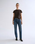 A model stands against a light gray studio background wearing a dark brown short-sleeve crewneck t-shirt, blue straight-leg jeans, and black ankle boots.