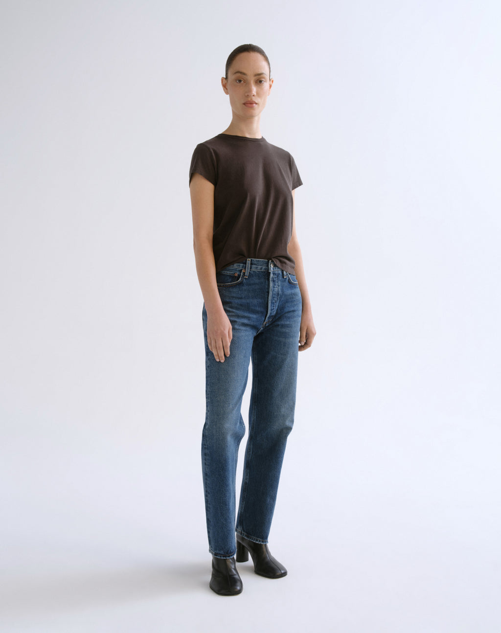 A model stands against a plain light gray studio background wearing a dark brown short-sleeve t-shirt, blue jeans, and black ankle boots.