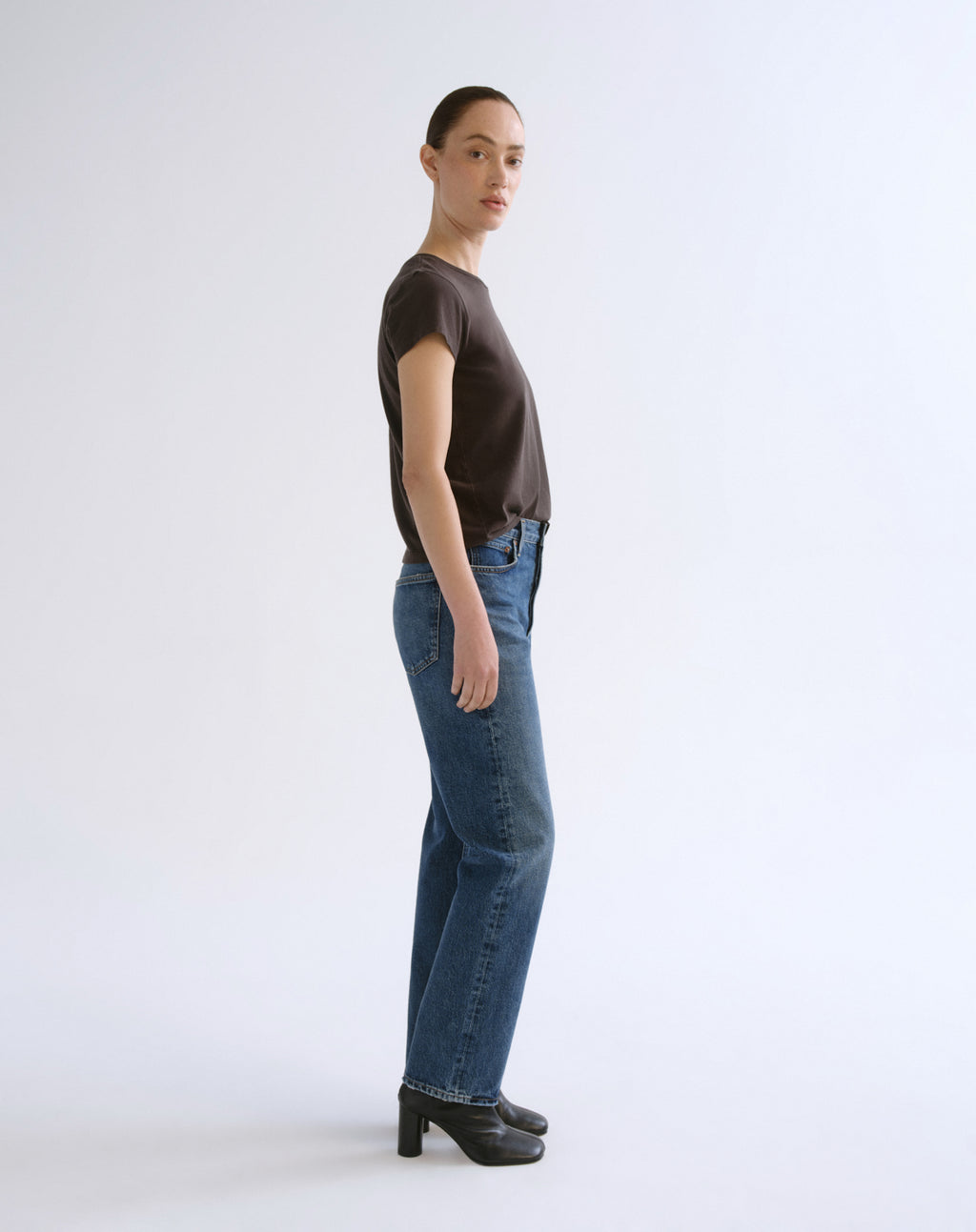 A model is shown in profile wearing a dark brown short-sleeve t-shirt, blue straight-leg jeans, and black high-heeled shoes against a plain white studio background.