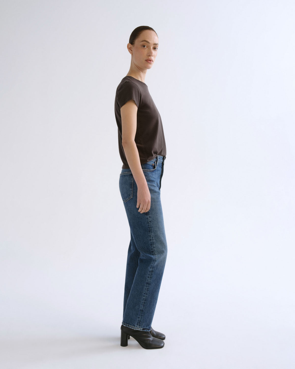 A model standing in profile wearing a dark brown short-sleeve crewneck t-shirt, blue straight-leg jeans, and black high-heeled shoes against a light gray studio background.