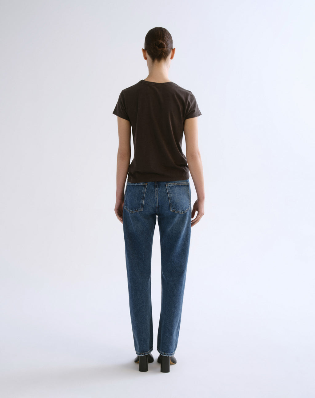 A model viewed from the back wearing a dark brown short-sleeve t-shirt and blue jeans, standing against a plain white studio background.