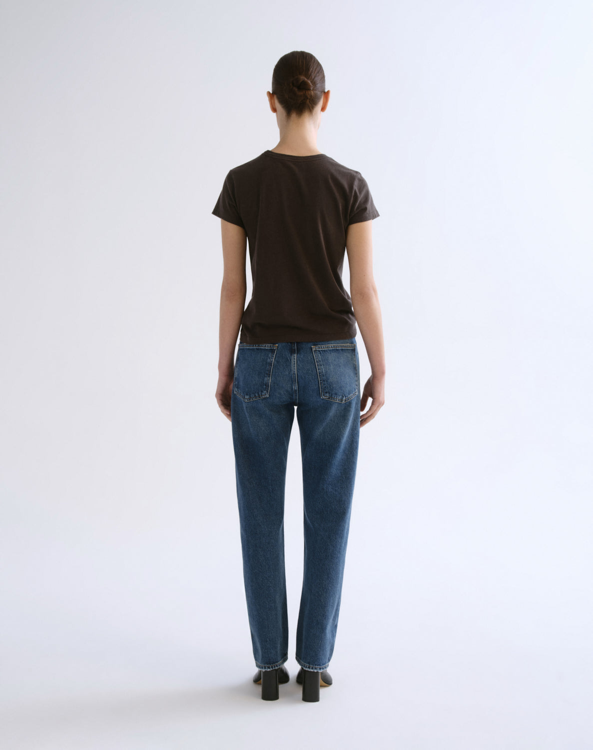 A model viewed from the back wearing a dark brown short-sleeve t-shirt and blue jeans, standing against a plain white studio background.