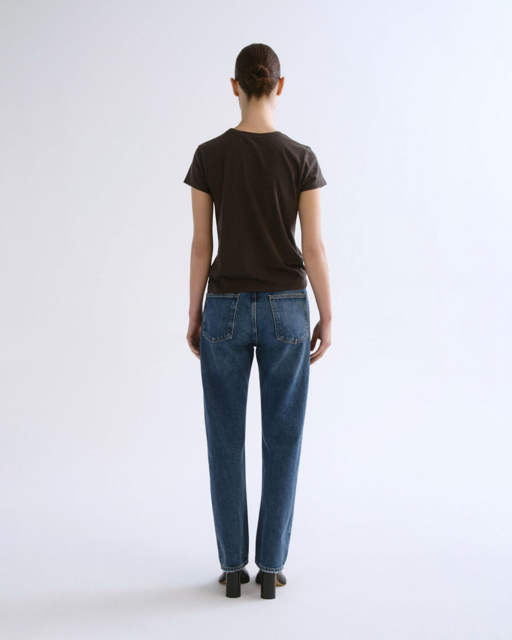 A model viewed from the back wearing a dark brown short-sleeve t-shirt and blue jeans, standing against a plain white studio background.