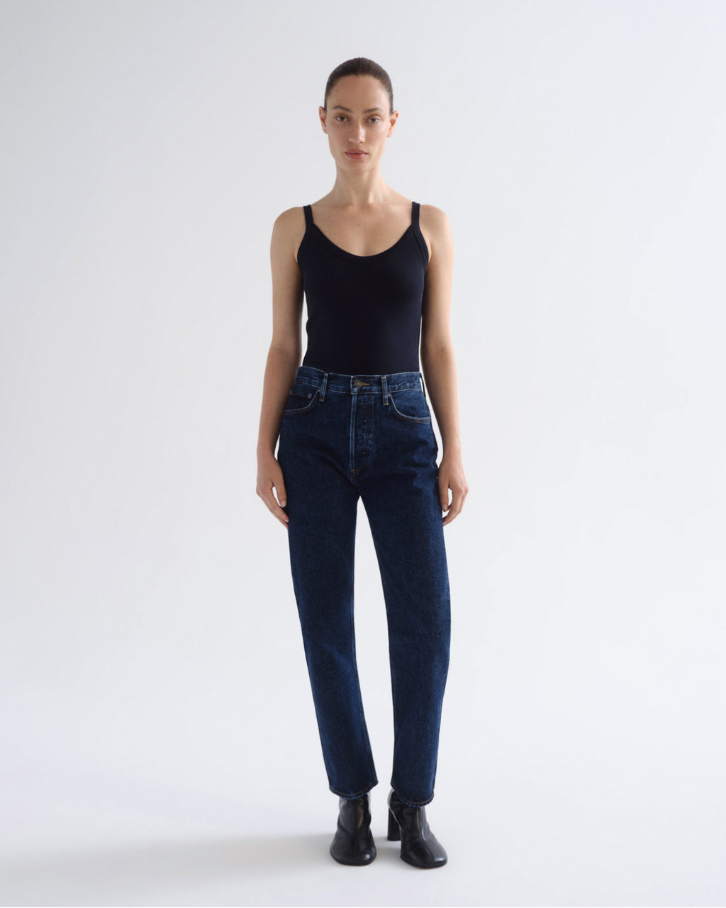 A model stands against a light gray studio background wearing a black ribbed tank top, dark blue high-waisted straight-leg jeans, and black ankle boots.