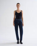 A model stands against a light gray studio background wearing a black ribbed tank top, dark blue high-waisted straight-leg jeans, and black ankle boots.